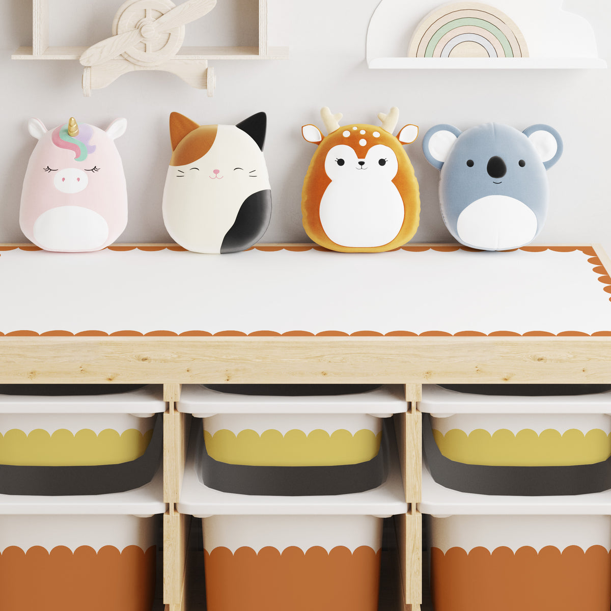 Four plush animal-shaped pillows or toys lined up on a light wood trofast unit the small drawer in scallop design, Yellow stone and the larger drawer in Rumba Orange. The characters are a pink unicorn, a calico cat, an orange-and-white deer, and a blue-gray koala, each with soft plush textures and cute facial features.