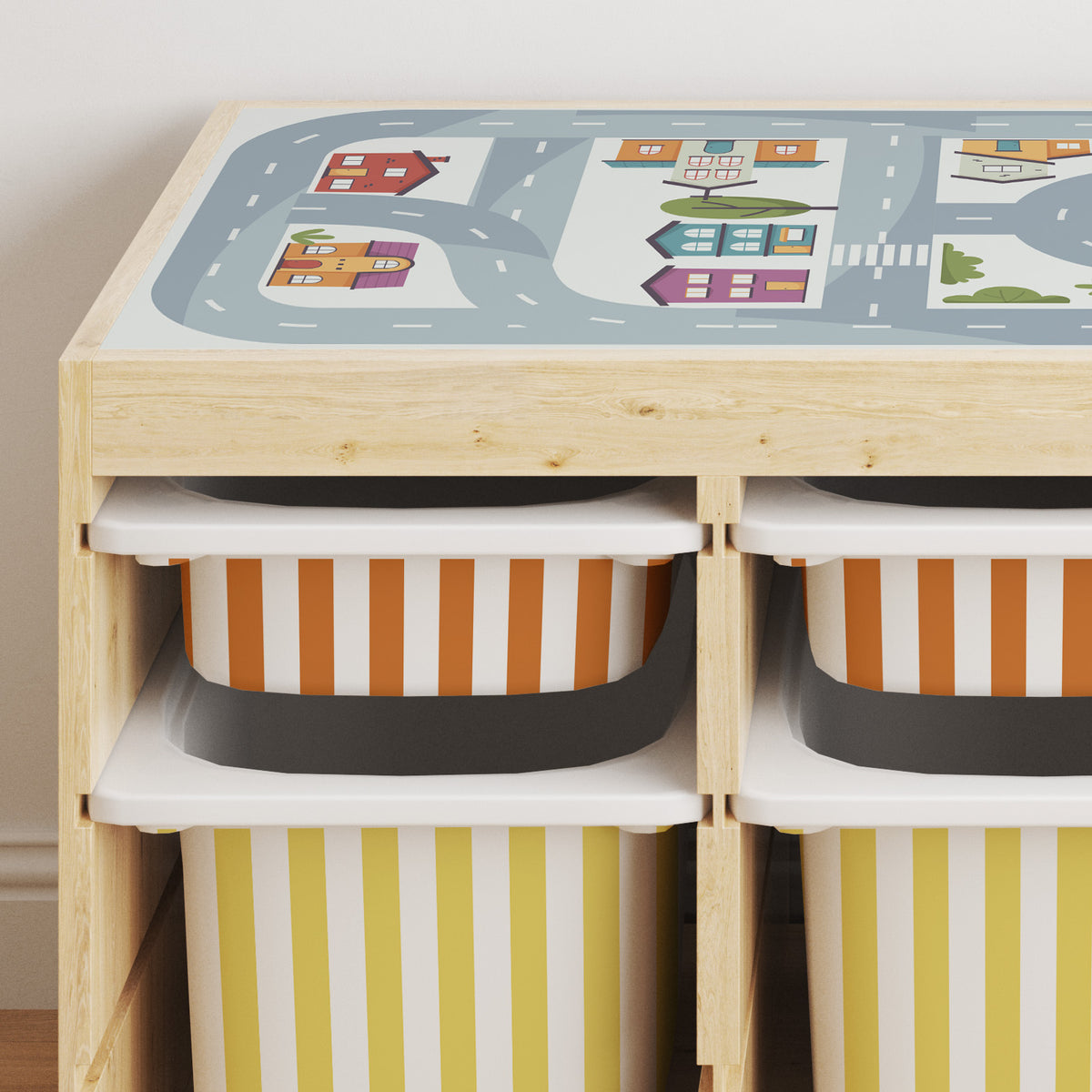 A light wood trofast storage cabinet with a colorful road-and-house play mat on top and four striped fabric bins in rumba orange, yellow stone, and white inside.