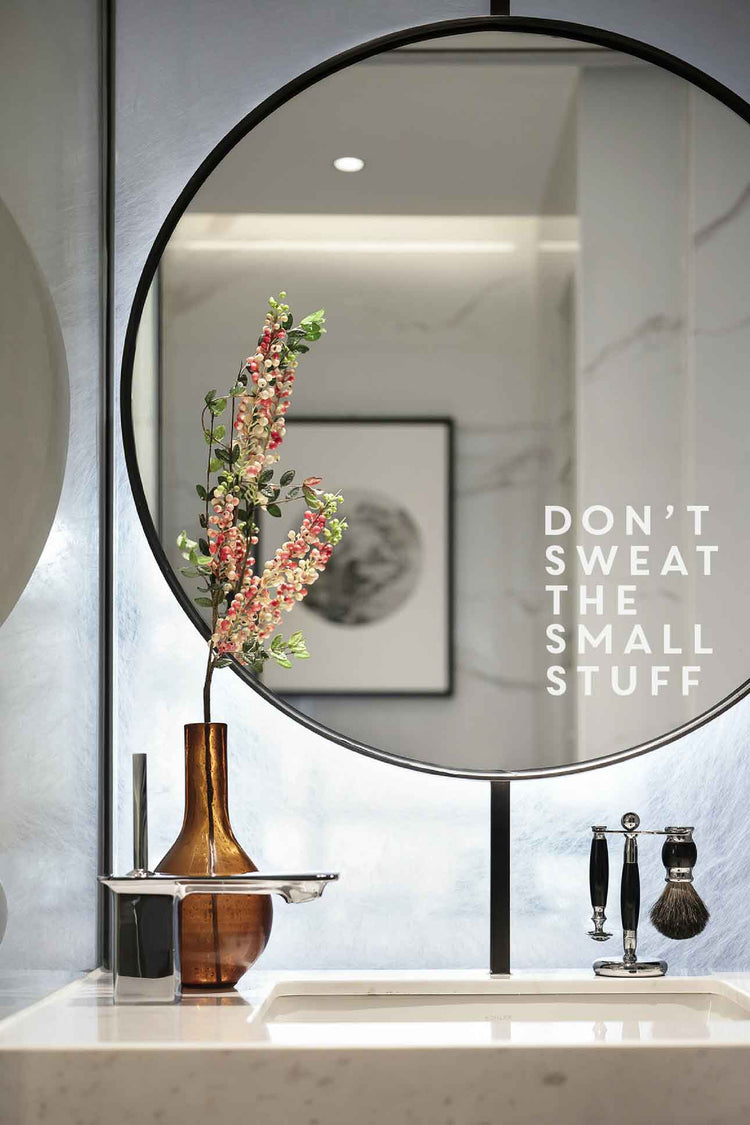 Round mirror on a wall with a vase of flowers and a motivational quote on a marble countertop.