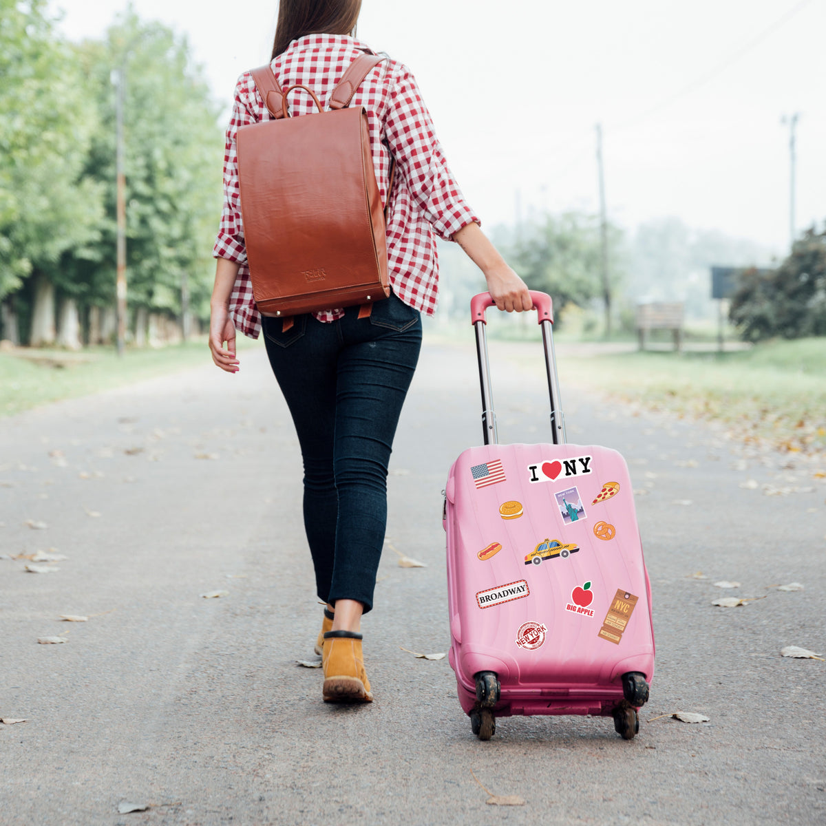 Person walking with a pink suitcase with New York Travel stckers and brown backpack on a path.