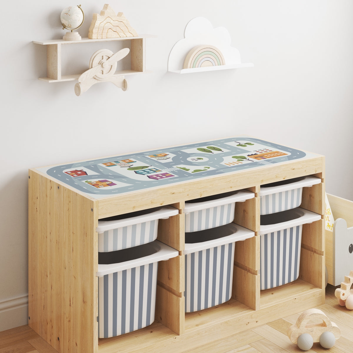 A light natural wood Trofast storage cabinet with six open cubbies small drawers in dusky blue and larger drawers in drizzle colour in a stripe pattern. The top surface is a blue mat printed with colorful illustrated vehicles and routes.