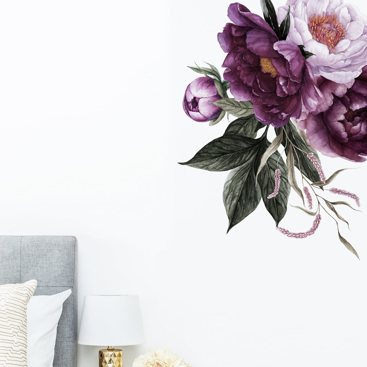 A wall sticker depicting a cluster of rich plum peonies applied on a white wall above a bed, with a lamp and pillows in the foreground.