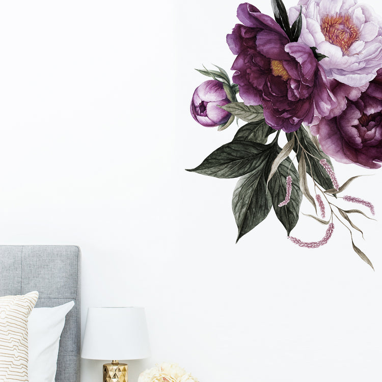 A wall sticker depicting a cluster of rich plum peonies applied on a white wall above a bed, with a lamp and pillows in the foreground.