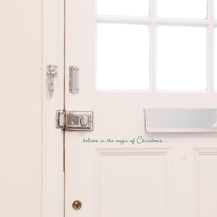Believe In The Magic of Christmas Front Door Sticker. Shown in a dark green colour on a white front door, in a script style font. A small Christmas decoration for a home.