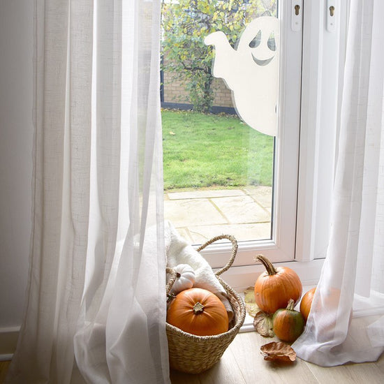 Cute white ghost sticker on some glass doors with pumpkins in a basket and arranged on the floor. The window has white curtains.