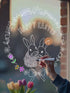 A person coloring in a window sticker with a marker, featuring an Easter-themed design with bunnies and flowers.