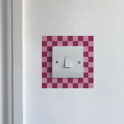 Light switch with a dark &amp; light pink checkered border on a white wall.