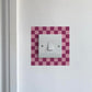 Light switch with a dark & light pink checkered border on a white wall.