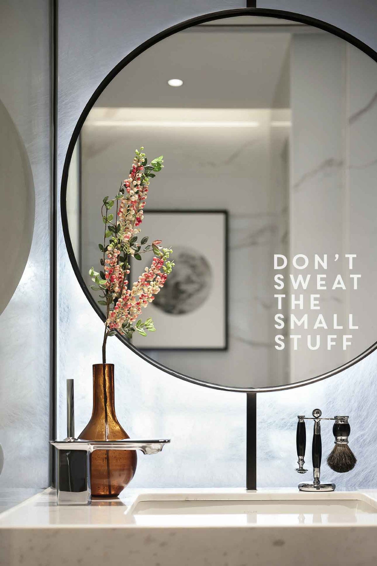 Round mirror on a wall with a vase of flowers and a motivational quote on a marble countertop.