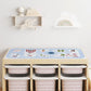A light wood toy storage unit with six pink patterned bins underneath, topped with a blue play mat featuring circus-themed illustrations. The unit is styled in a bright nursery setting with wooden wall shelves and toys above.