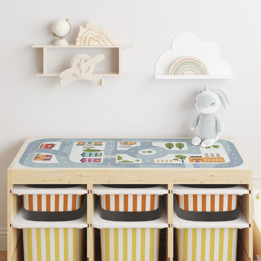 A light wood  trofast storage unit with small drawers in Rumba Orange and Large drawer in Yellow stoner with a play mat on top, three open cubbies with striped bins, a plush bunny on the side, and wall shelves above in a bright nursery setting.