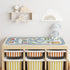 A light wood  trofast storage unit with small drawers in Rumba Orange and Large drawer in Yellow stoner with a play mat on top, three open cubbies with striped bins, a plush bunny on the side, and wall shelves above in a bright nursery setting.
