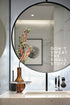 Round mirror on a wall with a vase of flowers and a motivational quote on a marble countertop.