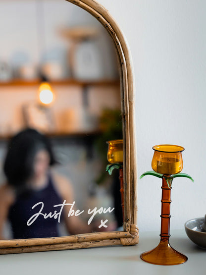 Amber candle holder on a surface with a mirror reflecting a blurred indoor setting.
