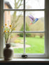 A vinyl hummingbird wall sticker and window decal applied to a window pane, with a vase of flowers on the windowsill and a view of a garden outside.