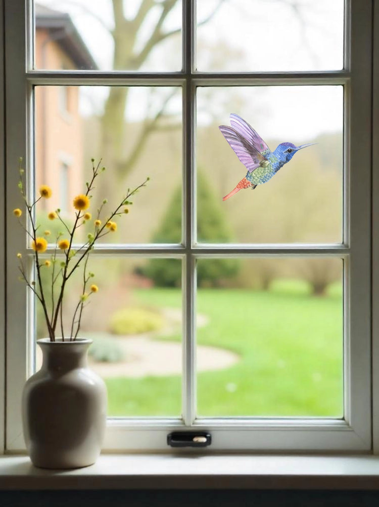 A vinyl hummingbird wall sticker and window decal applied to a window pane, with a vase of flowers on the windowsill and a view of a garden outside.