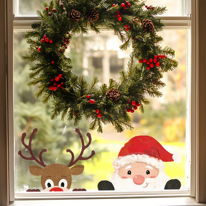 A festive Christmas window decoration featuring a brown reindeer head sticker with large antlers, a red nose, and white eyes, next to a santa head sticker with a red hat, white beard, black gloves and a cheeky face, positioned below a green pine garland with red berries.