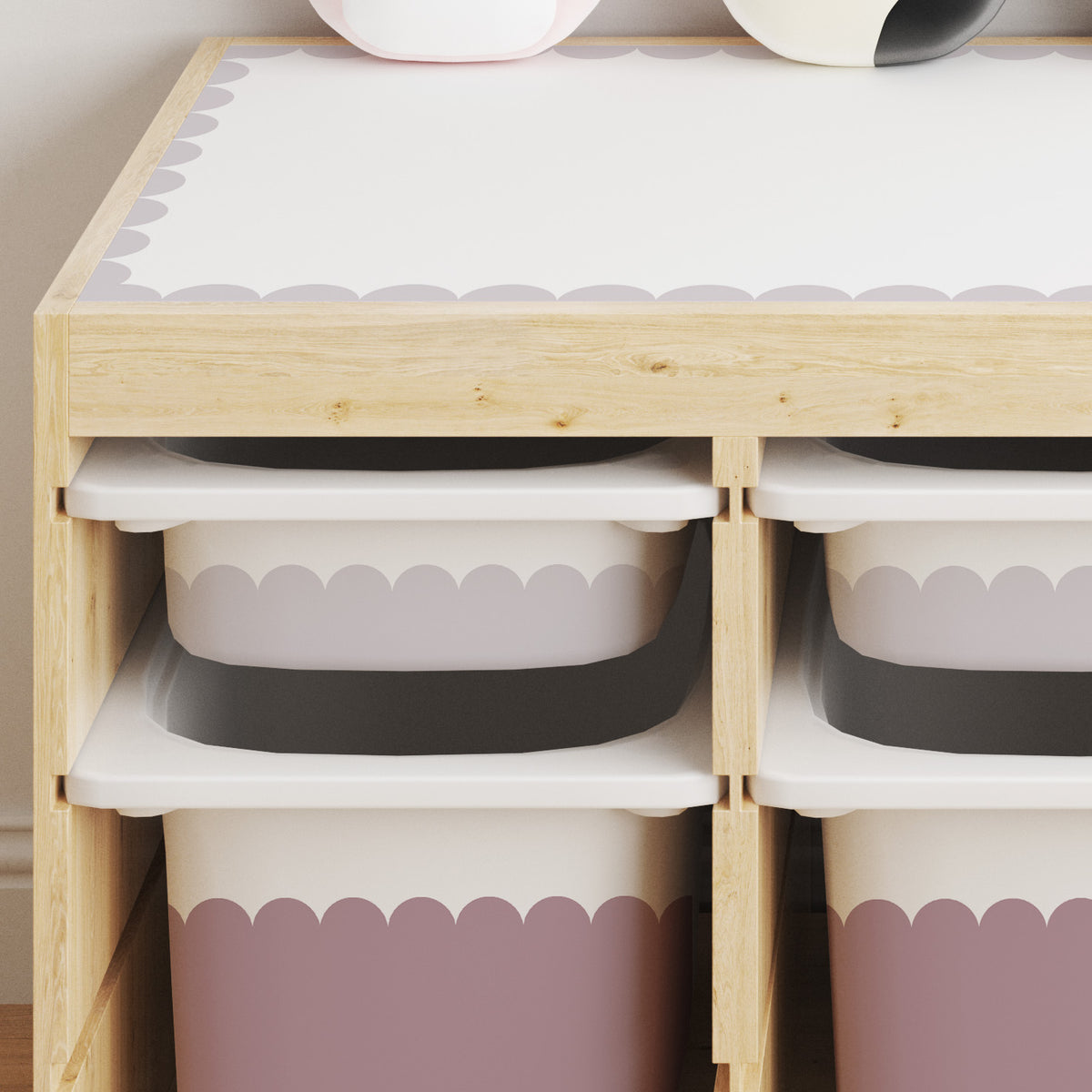 A light wood storage organizer with four open compartments holding white bins with scalloped stickers in Dusky Lilac and Rose Dusk, shown against a light beige wall.