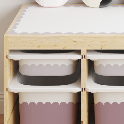 A light wood storage organizer with four open compartments holding white bins with scalloped stickers in Dusky Lilac and Rose Dusk, shown against a light beige wall.