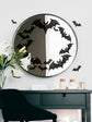 Round mirror with black bat decals on a white wall above a dark wooden desk.