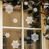 White lace snowflake window decals applied to a multi-pane window, with a holiday garland and warm string lights visible in the background.
