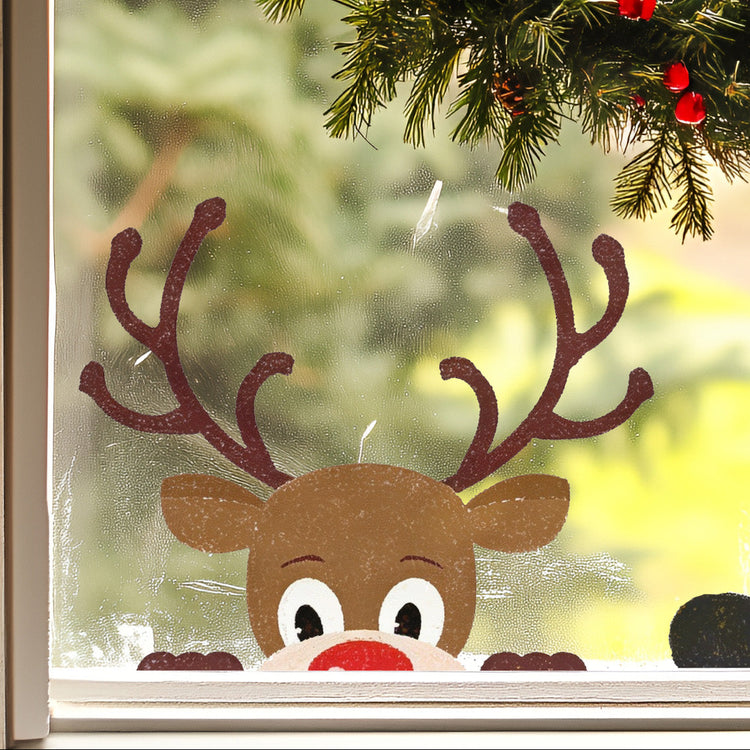 A festive Christmas window decoration featuring a brown reindeer head sticker with large antlers, a red nose, and white eyes, positioned below a green pine garland with red berries.