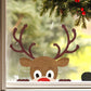 A festive Christmas window decoration featuring a brown reindeer head sticker with large antlers, a red nose, and white eyes, positioned below a green pine garland with red berries.