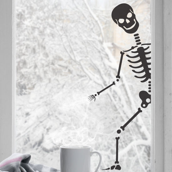 A black colored peeping skeleton sticker shown peeping through a window, with a cup of coffee and a blanket in the foreground.