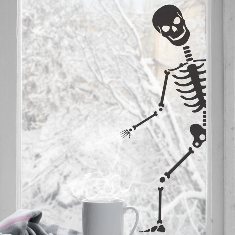 A black colored peeping skeleton sticker shown peeping through a window, with a cup of coffee and a blanket in the foreground.