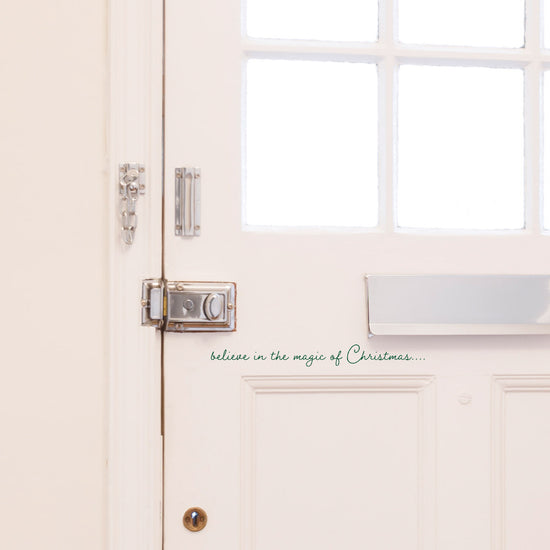 Believe In The Magic of Christmas Front Door Sticker. Shown in a dark green colour on a white front door, in a script style font. A small Christmas decoration for a home. 